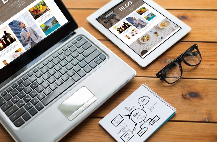 media, internet, business and technology concept - close up of blog web page on laptop computer screen, tablet pc, notebook and eyeglasses on wooden table